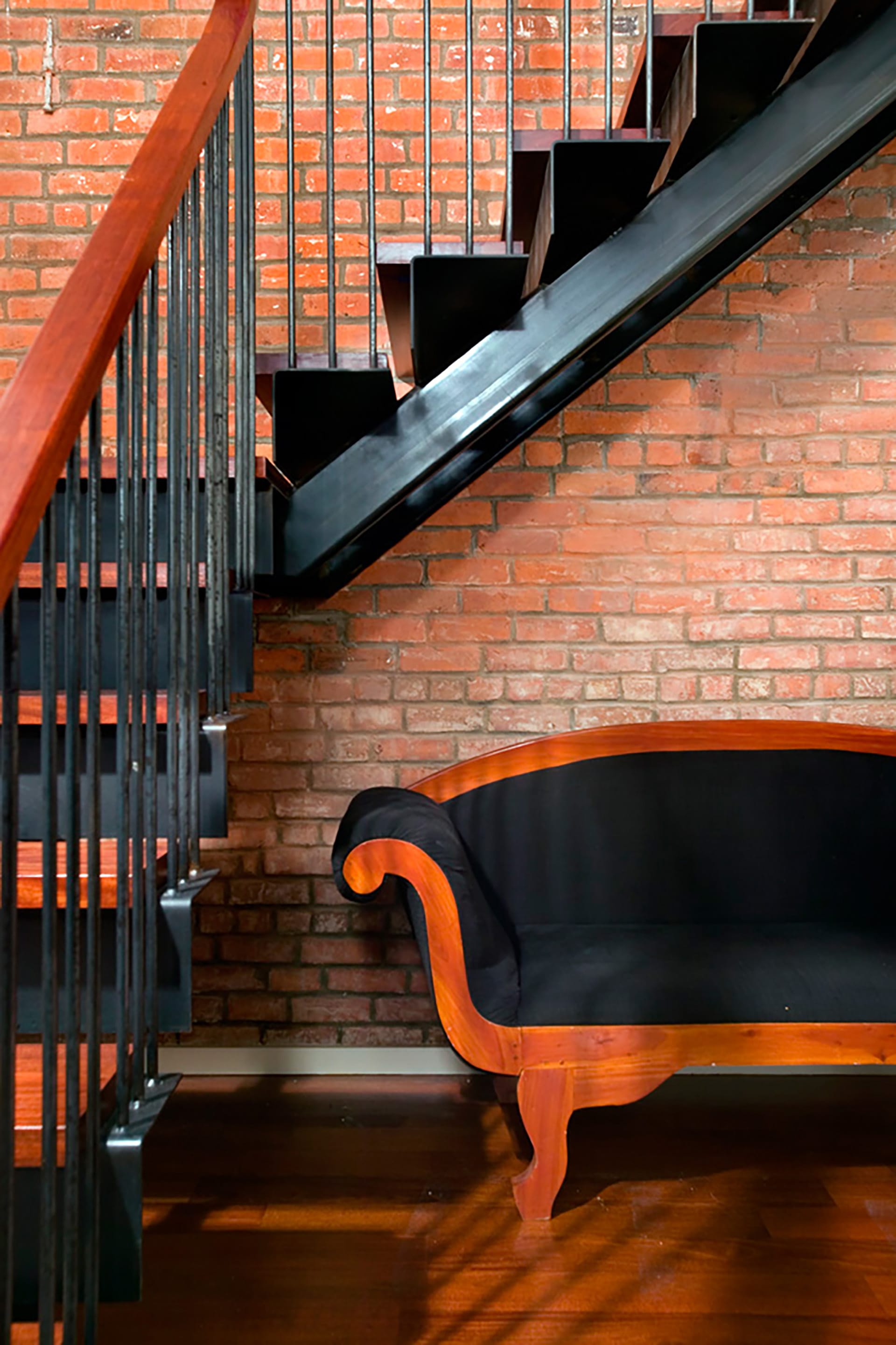 A staircase with black metal center beam stringers, cherry handrails and risers, and black balusters sits in front of an exposed brick wall. A wood and black fabric loveseat sits under the staircase.