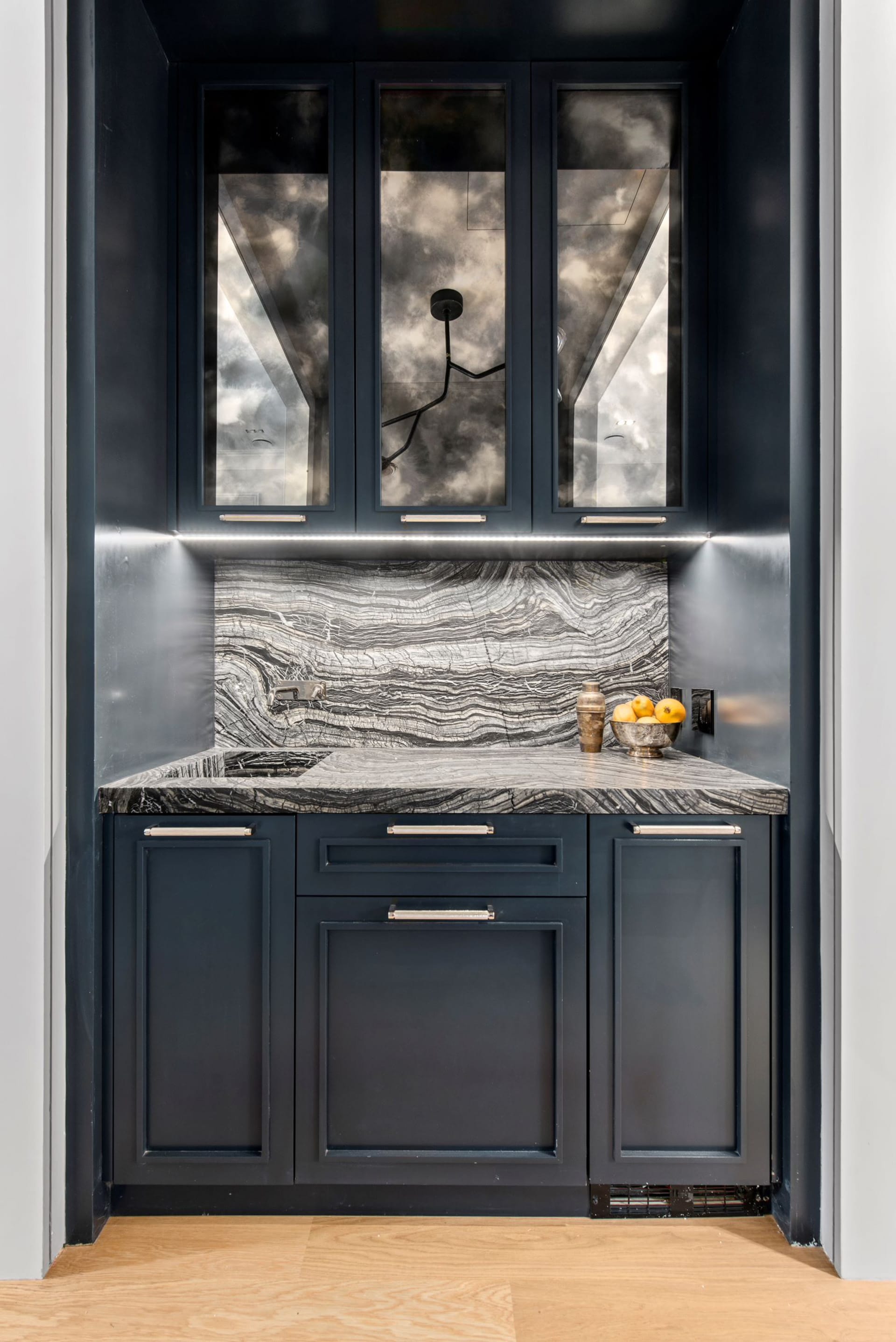 Wet bar with dark blue millwork, black and white striped stone countertop and backsplash, and light wood floors.