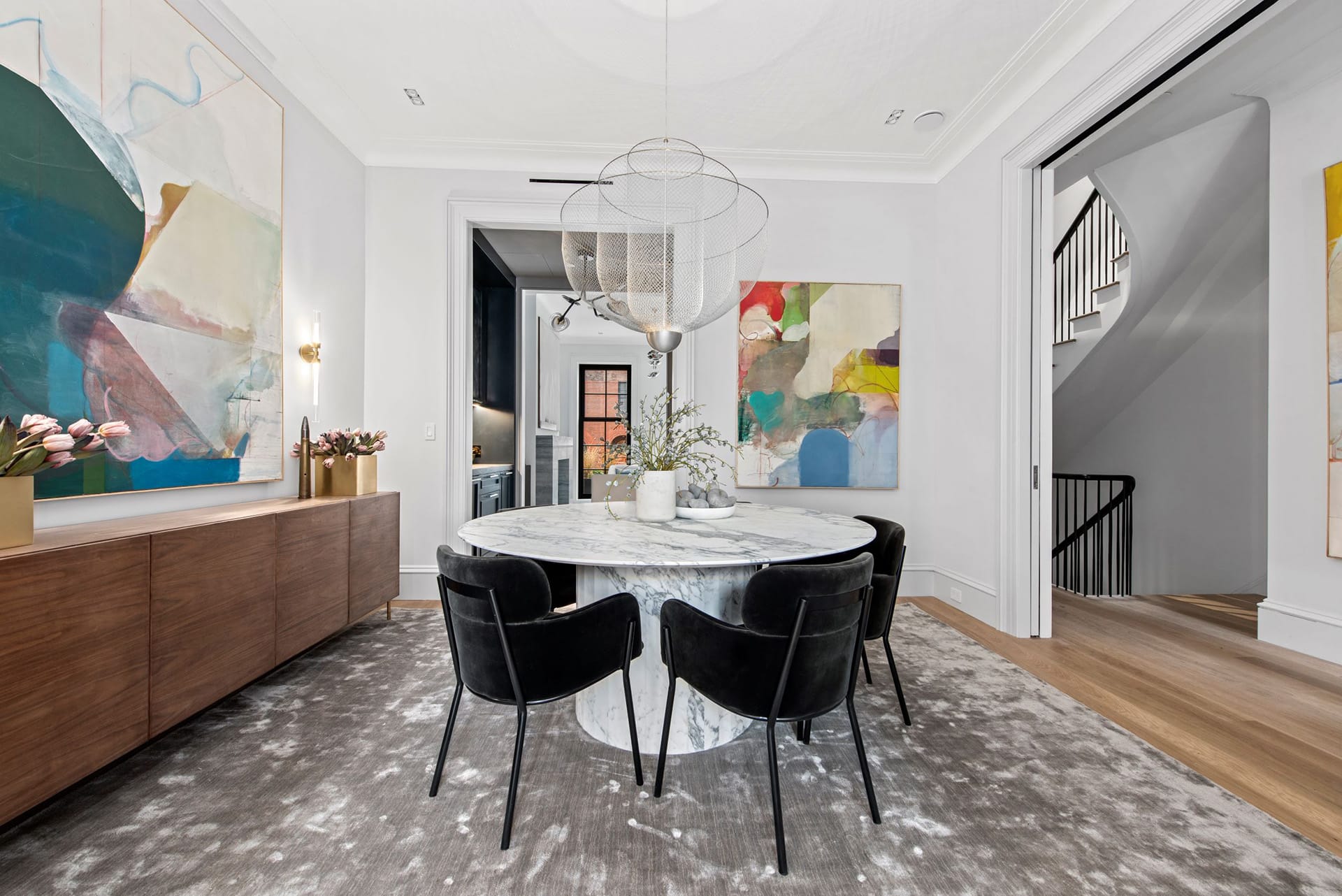 Dining room with a large grey area rug, marble dining table, black velvet dining chairs, wood credenza, two large paintings hanging on the walls, and a large silver chandlier.