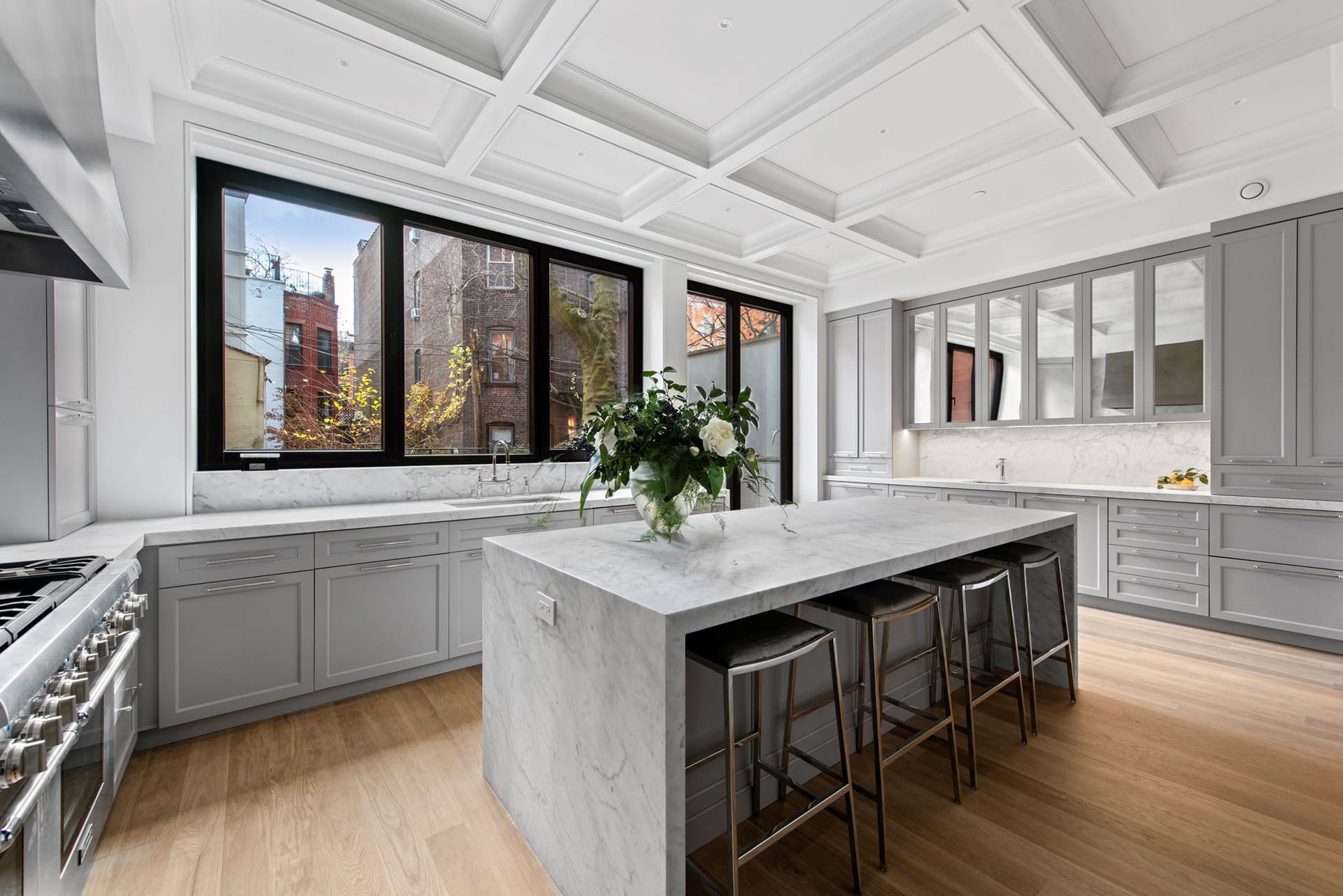 Full-width kitchen in a Brooklyn Heights townhouse with light grey millwork surrounded by marble countertops, island, and backsplash. Four barstools are lined up at the built-in eating area on the island. The high ceilings are covered in paneling.