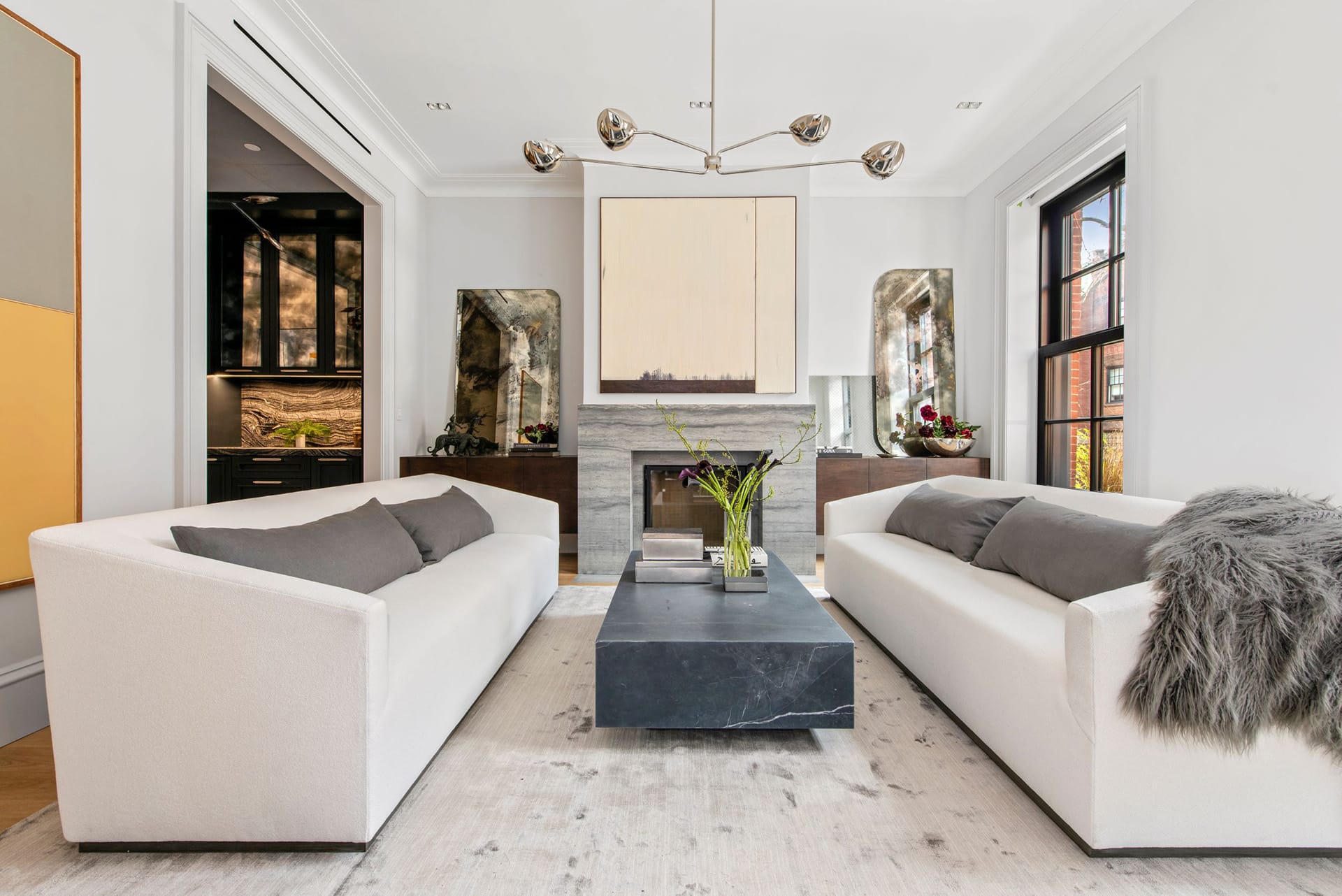 Living room with two large white couches facing each other. A large marble coffee table sits in between them, in front of a large, modern, marble mantel. Above the mantel is a large mirror. Above the coffee table, a large chrome chandelier hangs. On either side of the mantel, credenzas with aged glass mirrors provide storage.