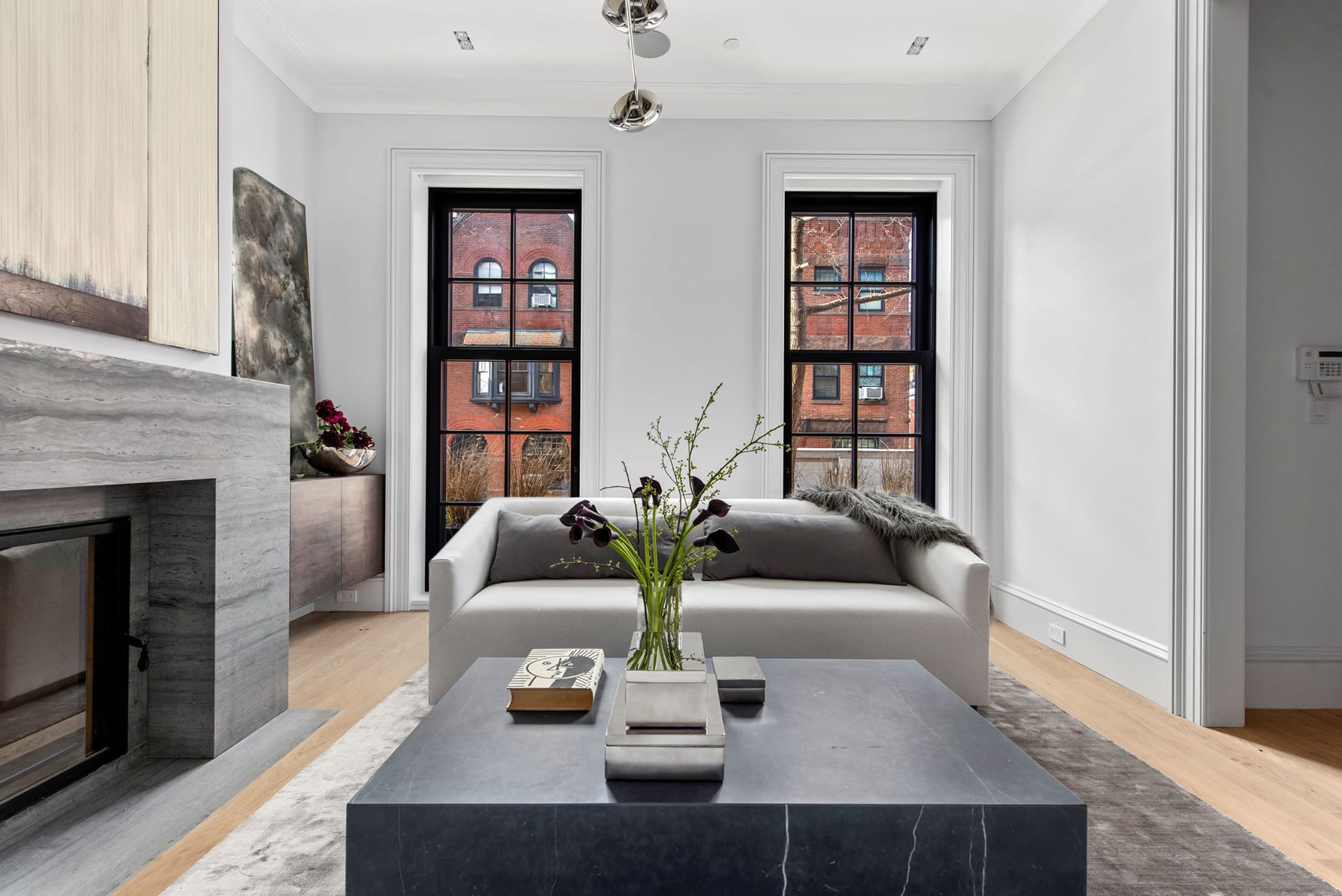 Living room with a large marble coffee table and white couch, in front of a large, modern, marble mantel. Above the mantel is a large mirror. Above the coffee table, a large chrome chandelier hangs. On either side of the mantel, credenzas with aged glass mirrors provide storage.