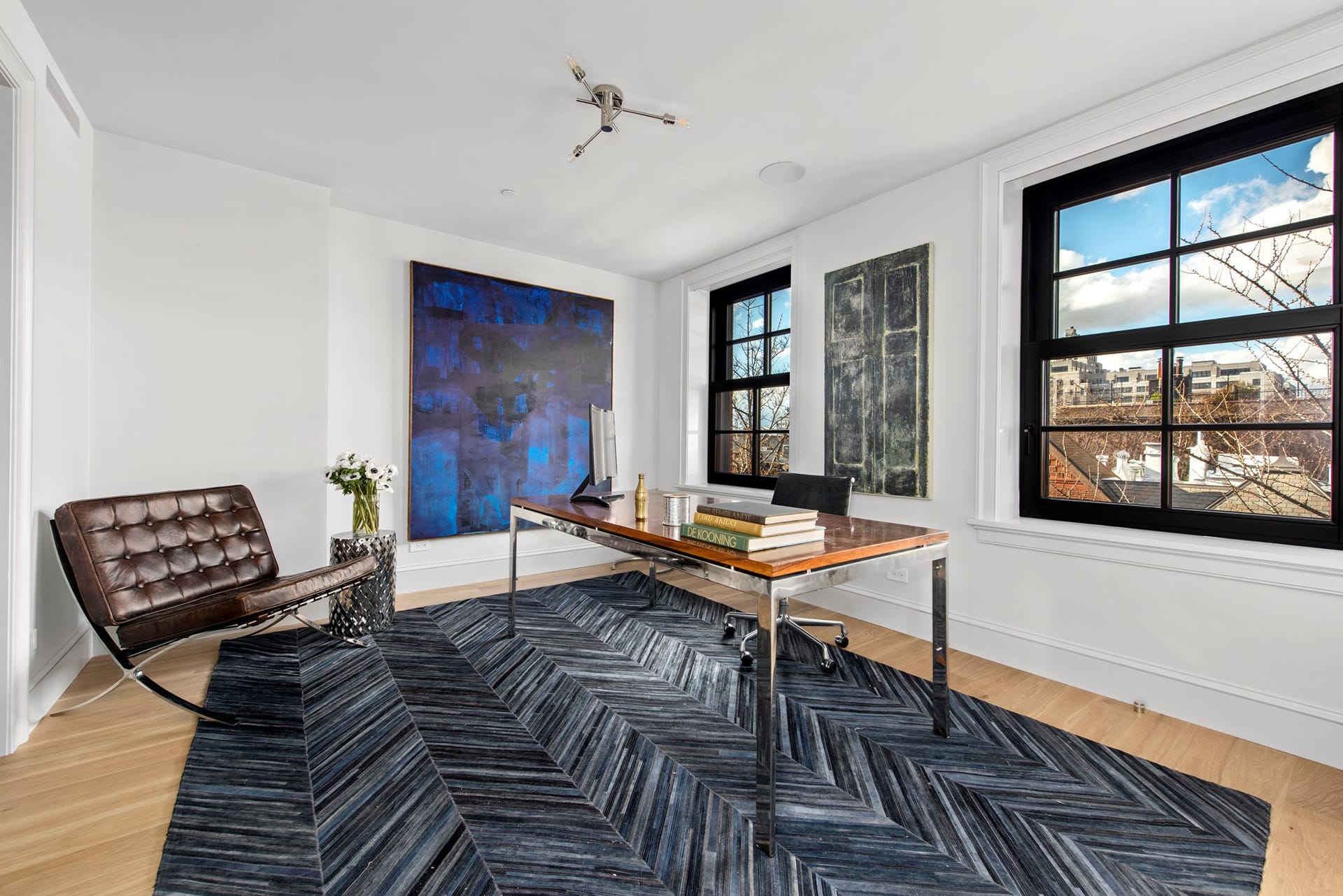Office with a chrome and lacquered wood desk, office chair, and a guest chair next to it. Two large paintings hang on the walls, and a large blue chevron area rug covers the white oak floors.