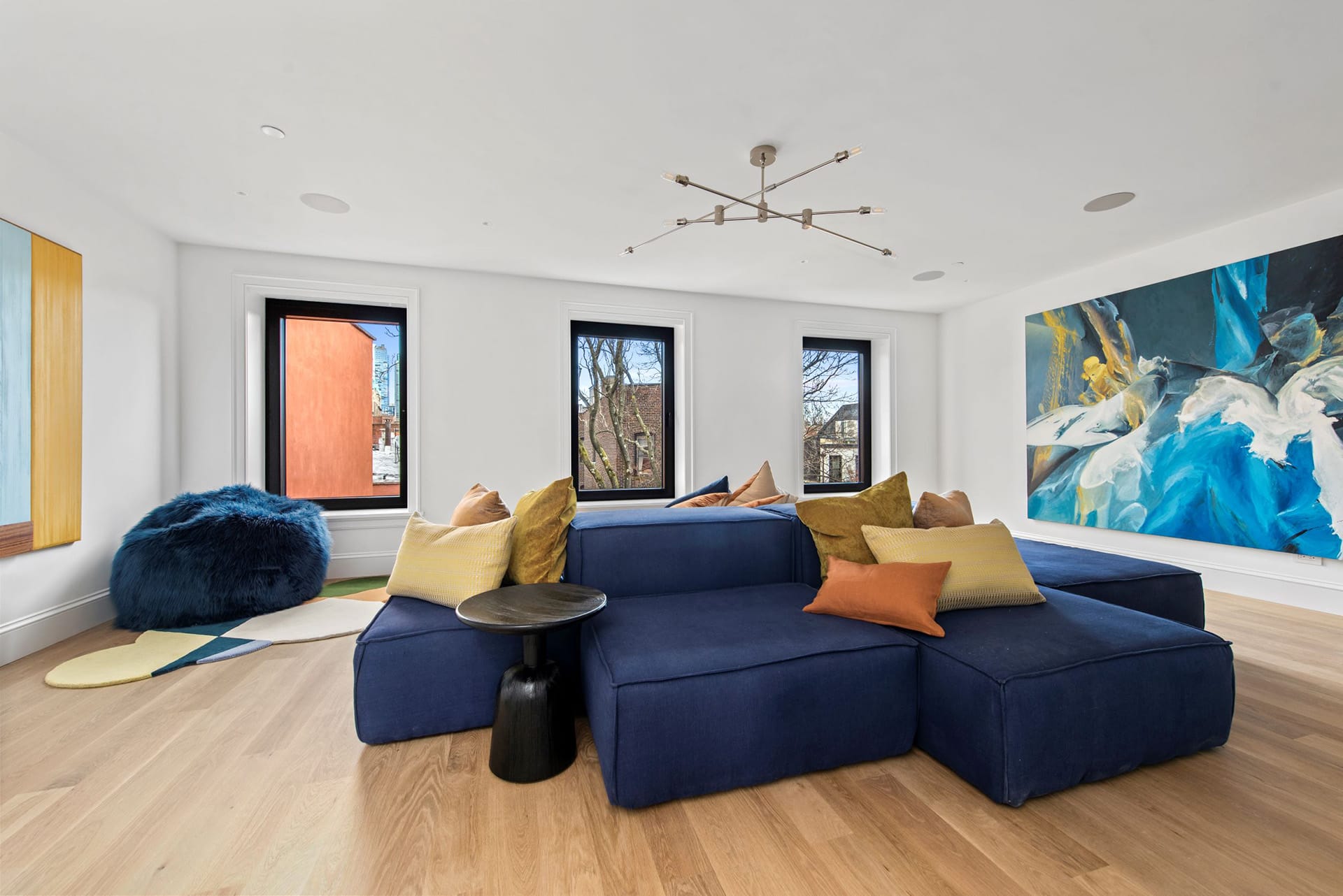 Recreation room with wood floors, three windows, and a large navy blue sectional with orange and yellow pillows.