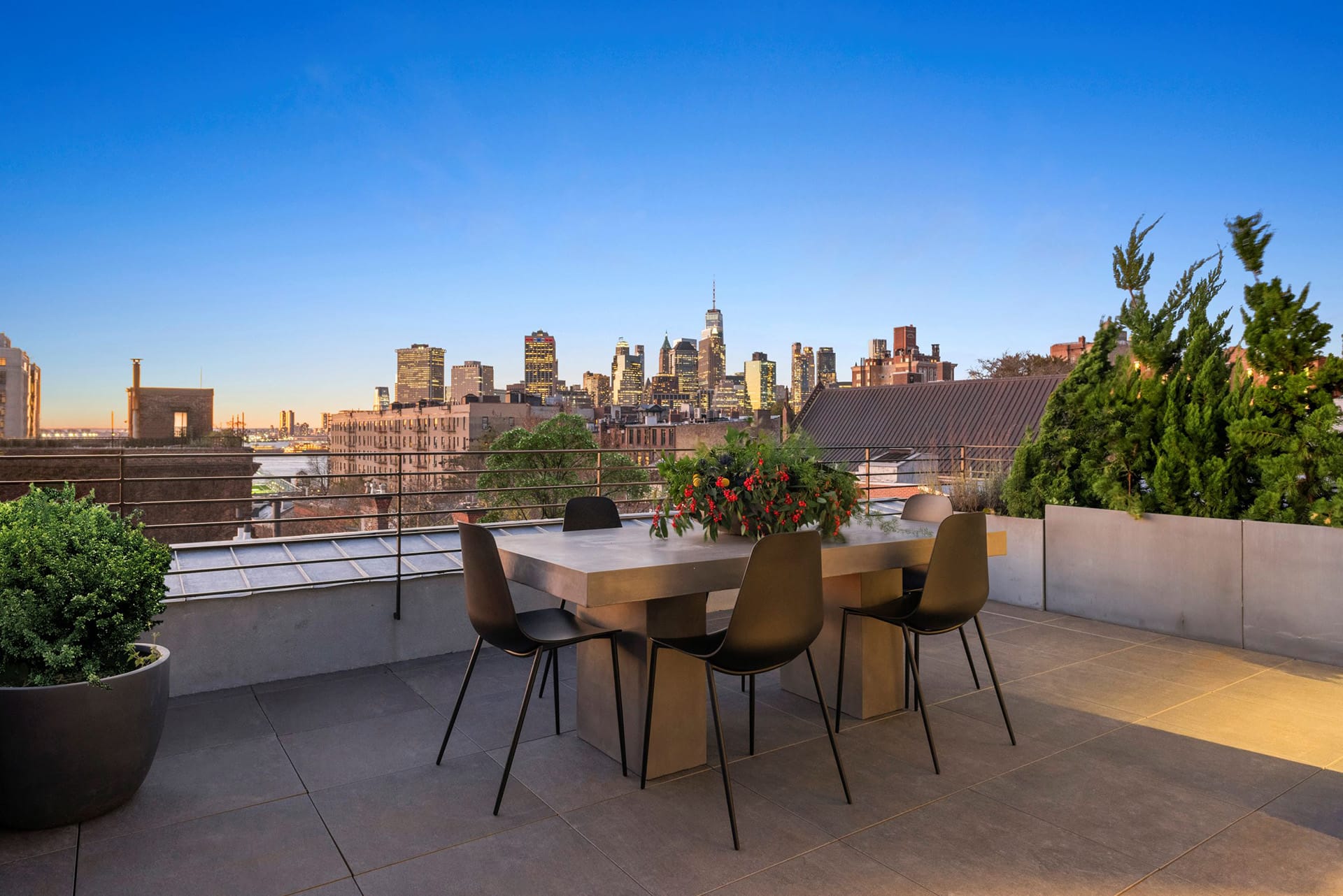 Rooftop with bluestone pavers, a dining table and chairs, lush evergreen landscaping, and sweeping views of the Manhattan skyline.