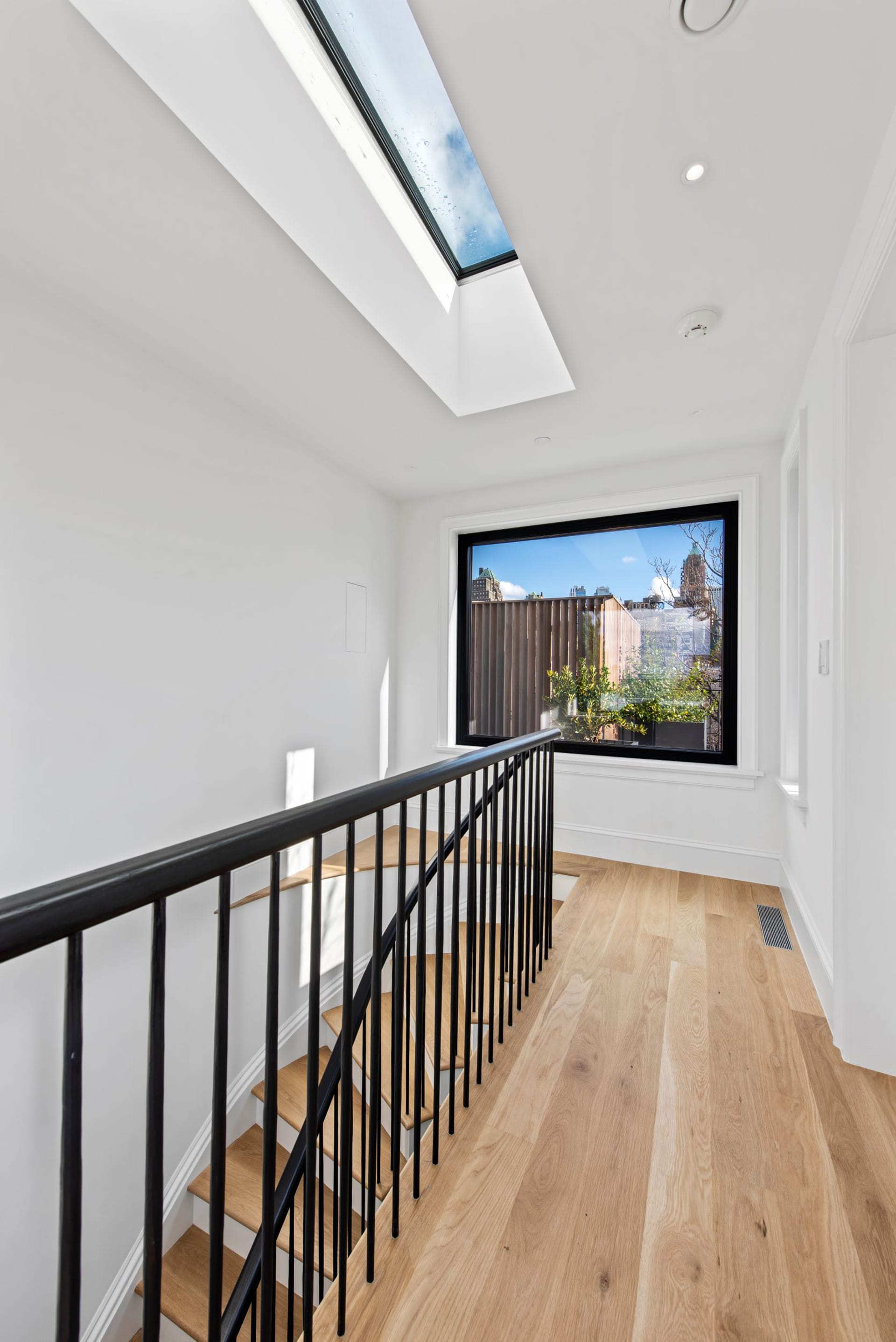 Fifth floor landing of a five-story townhouse with a large skylight above the staircase, and a large window at the landing. The floors are white oak and the walls are painted white.