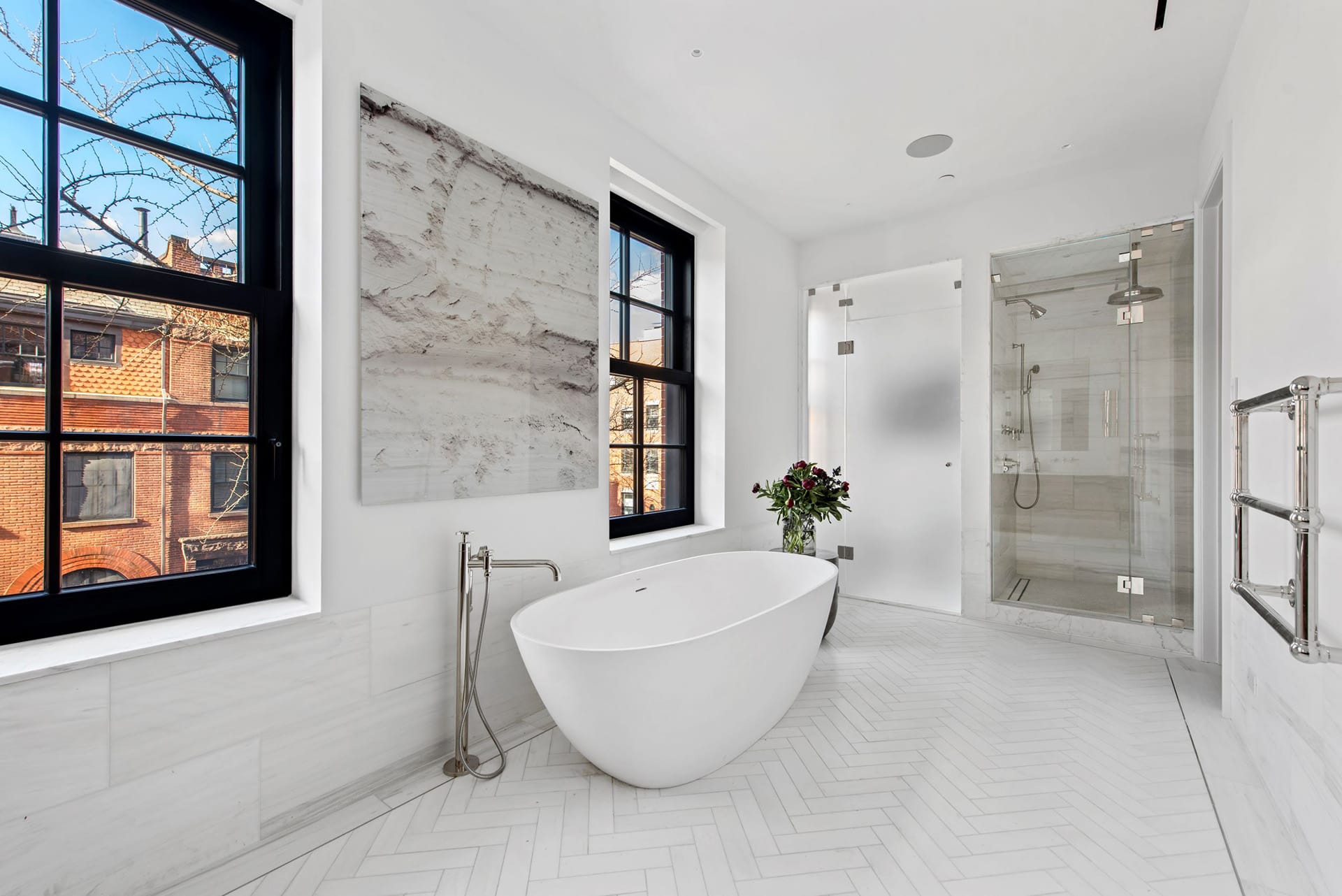 Primary bath with a freestanding tub in between two windows, a separate shower stall, heated towel bar, and white tile flooring.