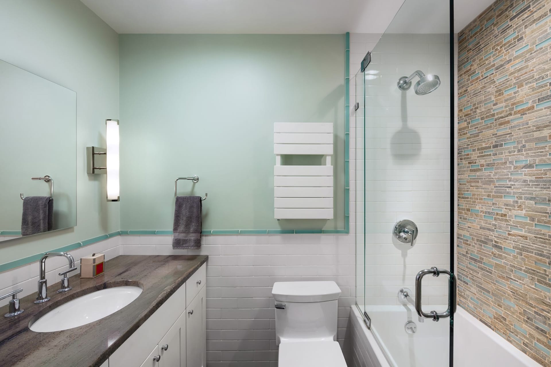 Primary bathroom with light teal walls, glass tiles in the shower, and natural stone countertops