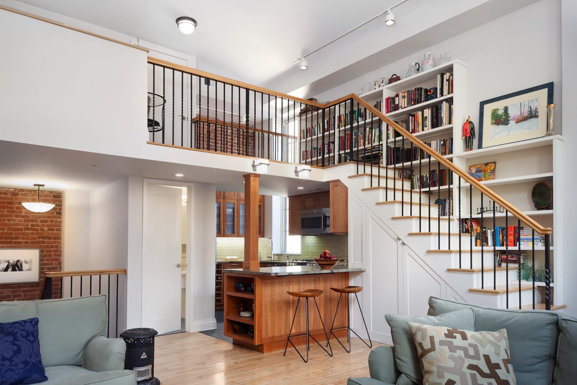Main living space of a loft apartment with an open kitchen, staircase lined with bookshelves, and metal railings opening the loft space to below.