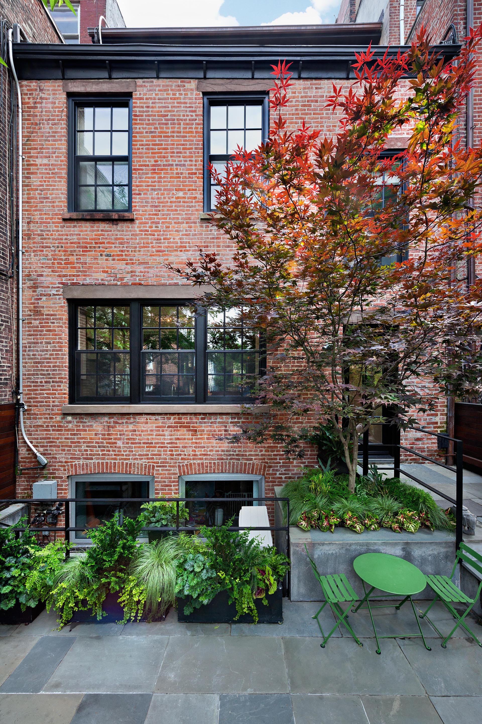 Rear façade of a brick house with a red tree, bluestone pavers, and green table and chairs