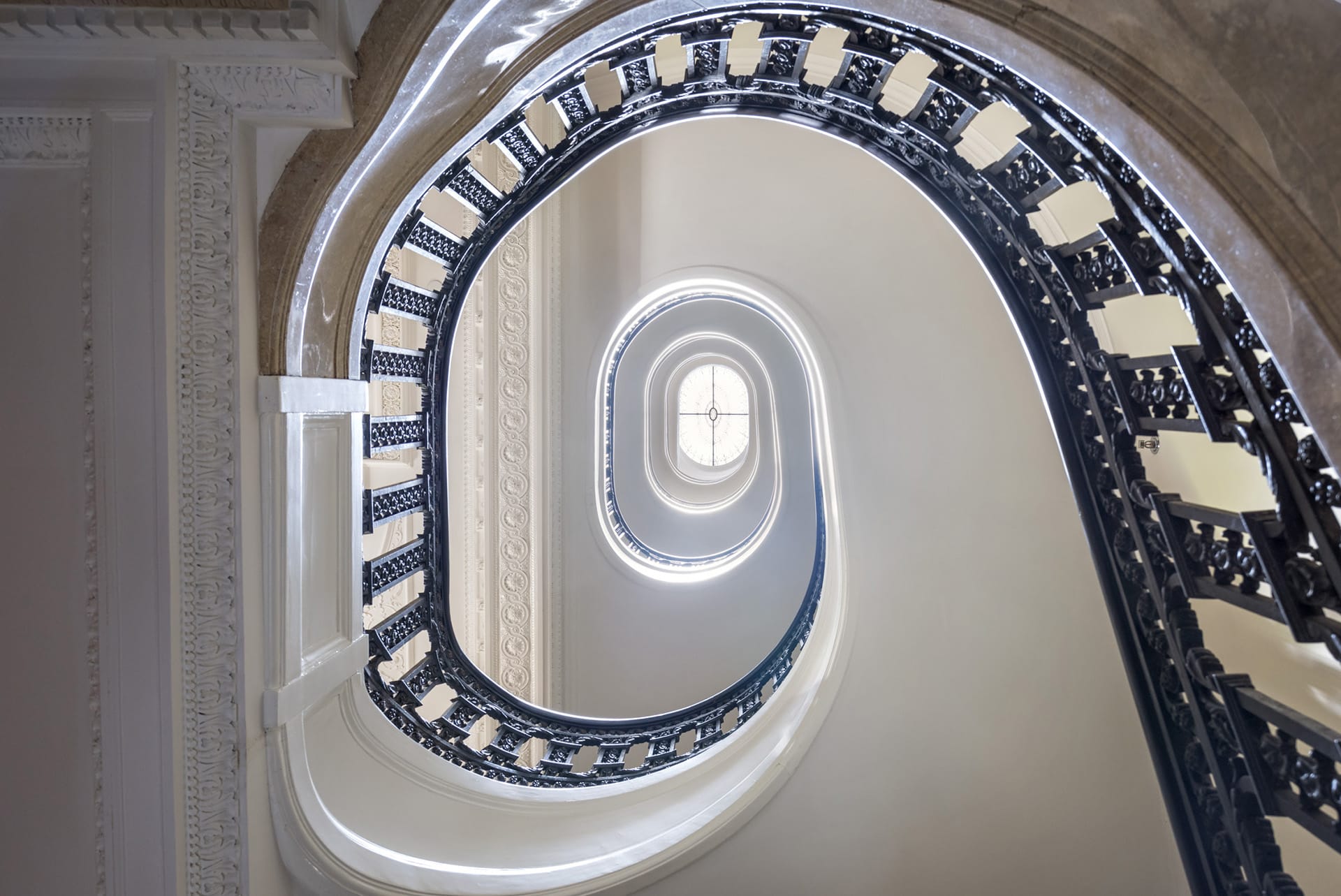 View from the first floor landing of a historic spiral staircase with intricate iron railings and a stained glass skylight.