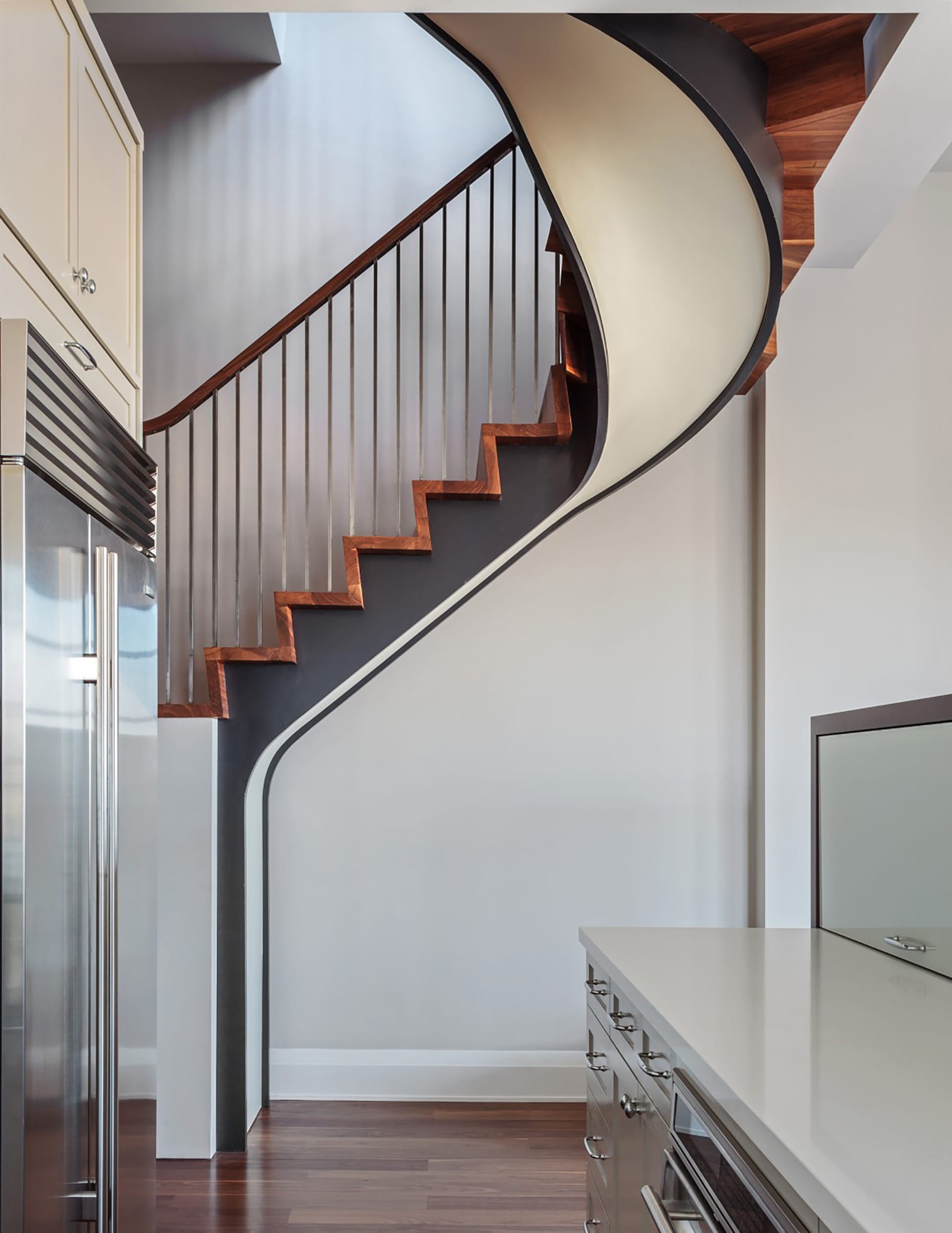 Sculptural staircase with black handrail and balusters, wood risers, and a white underside, next to the kitchen
