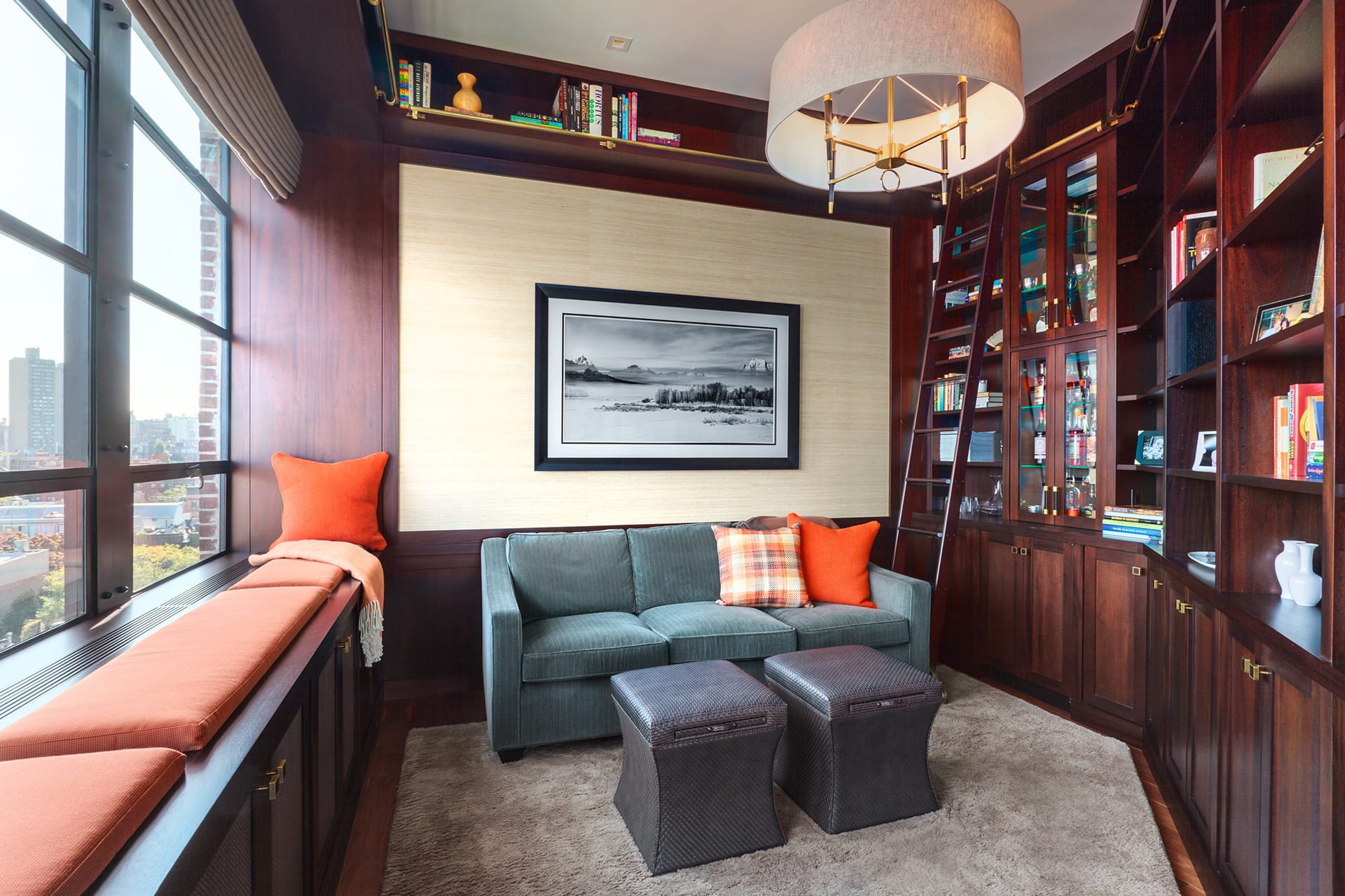 Library in a West Village townhouse. The room is wrapped in built-in dark wood bookshelves and millwork. A teal loveseat sits next to the window.