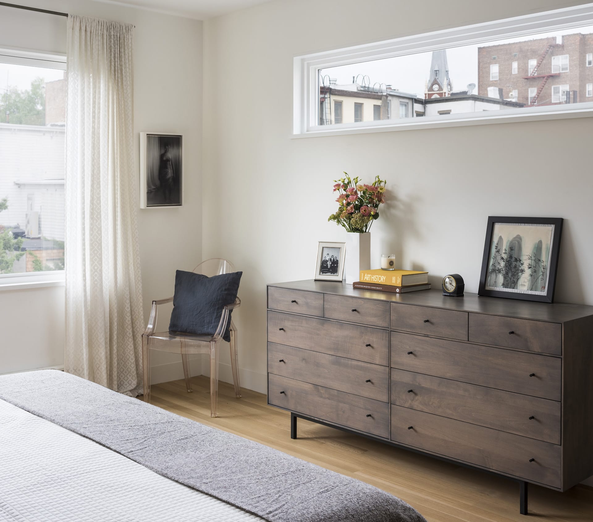 Bedroom with dark wood dresser, clear chair in the corner, and long, skinny window above the dresser