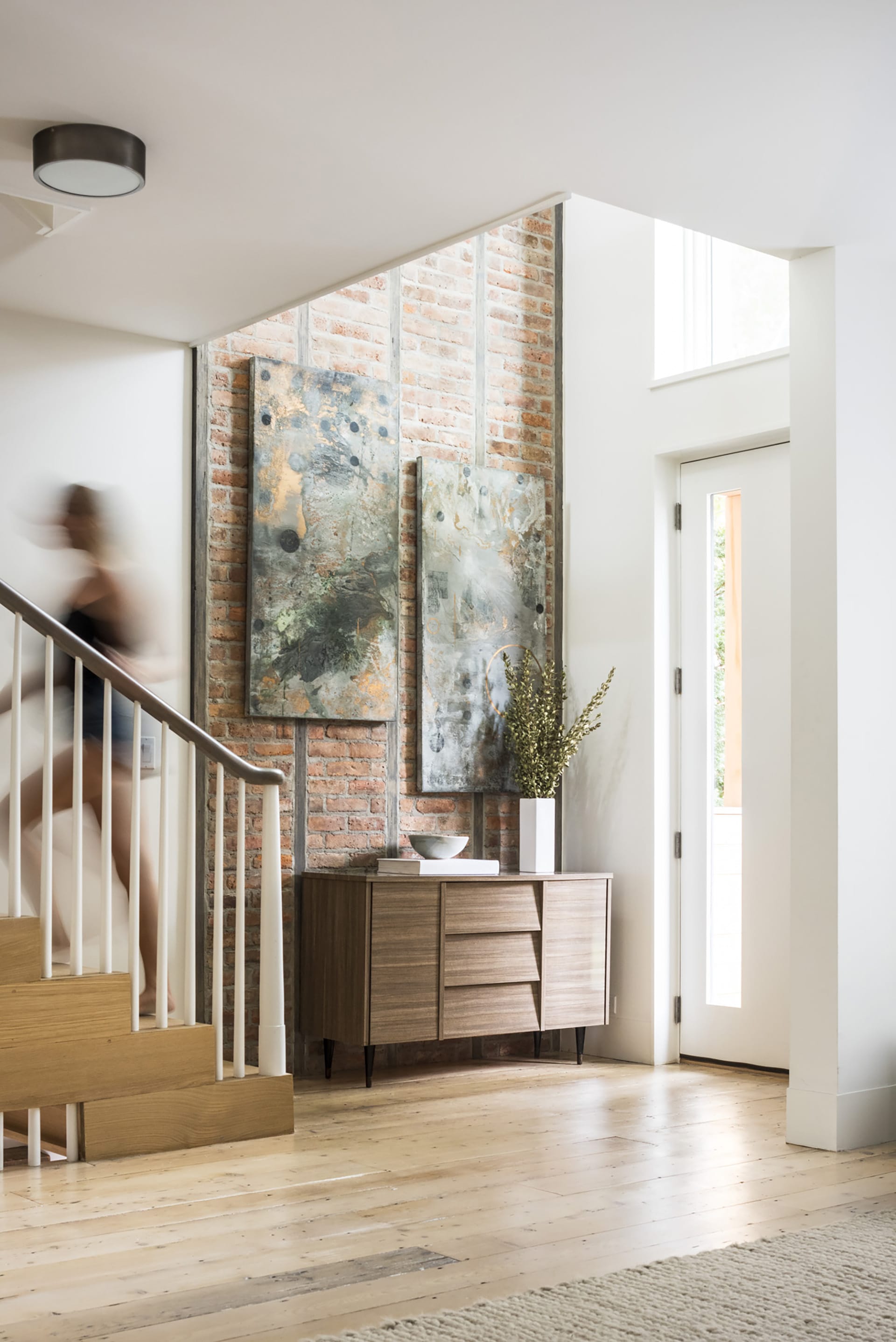 Foyer after renovation, with exposed brick wall, double height space, refurbished wood floors, and a woman walking up the stairs to the left