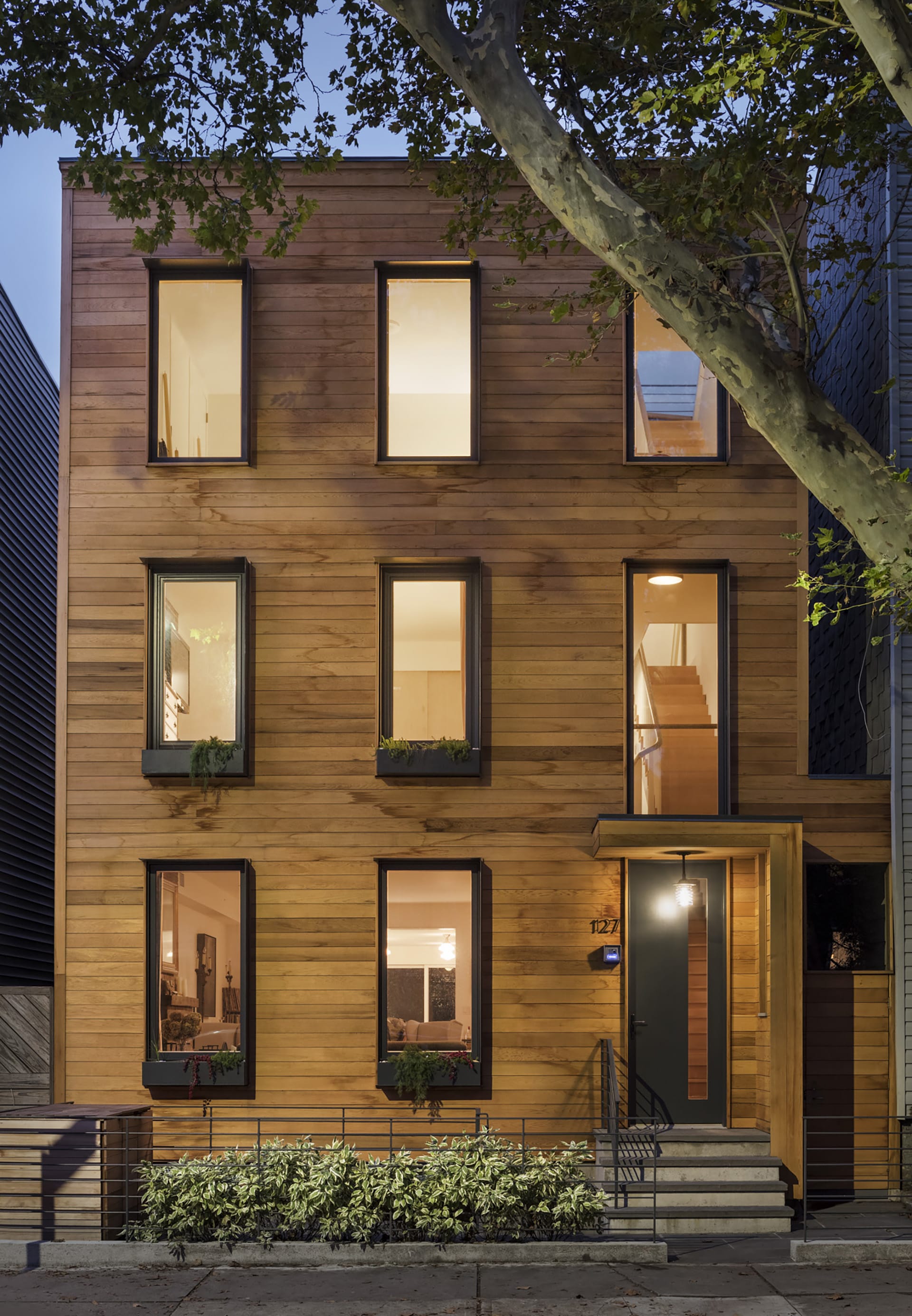 Exterior of a wood-framed Greenpoint townhouse with cedar siding, planters in front of the home and windows, and black window and door openings at night.