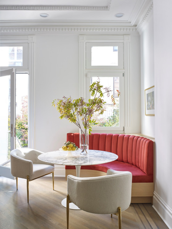 Corner of a kitchen with white walls and crown molding. A corner banquette is covered with bright coral red fabric. A marble kitchen table with plants on top of it and two white chairs sit in front of the banquette.