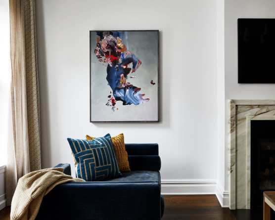 A grey abstract painting hangs on a white wall with a blue velvet tête-à-tête in front of it. To the left of the photo, tan curtains stretch from the top of a window to the floor. To the right of the frame, a tv hangs above a fireplace and marble mantle.